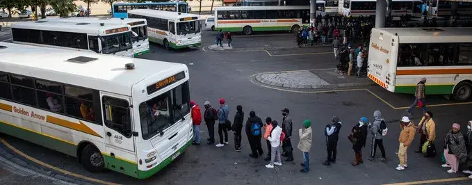 Blick über einen großen Busbahnhof in Kapstadt. Mehrere Busse stehen nebeneinander an den Haltestellen. Zwischen den Fahrzeugen und entlang der überdachten Bahnsteige sind zahlreiche Menschen unterwegs oder warten auf ihre Abfahrt. Im Hintergrund erstreckt sich die dichte Bebauung der Stadt; dahinter erhebt sich die Berglandschaft Kapstadts.