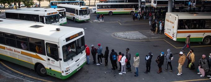 Blick über einen großen Busbahnhof in Kapstadt. Mehrere Busse stehen nebeneinander an den Haltestellen. Zwischen den Fahrzeugen und entlang der überdachten Bahnsteige sind zahlreiche Menschen unterwegs oder warten auf ihre Abfahrt. Im Hintergrund erstreckt sich die dichte Bebauung der Stadt; dahinter erhebt sich die Berglandschaft Kapstadts.