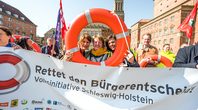 Im Fokus sind drei Frauen, welche durch einen Rettungsring gucken, vor ihnen ist ein Transparent mit der Aufschrift "Rettet den Bürgerentscheid!, Absatz Volksinitiative Schleswig-Holstein".