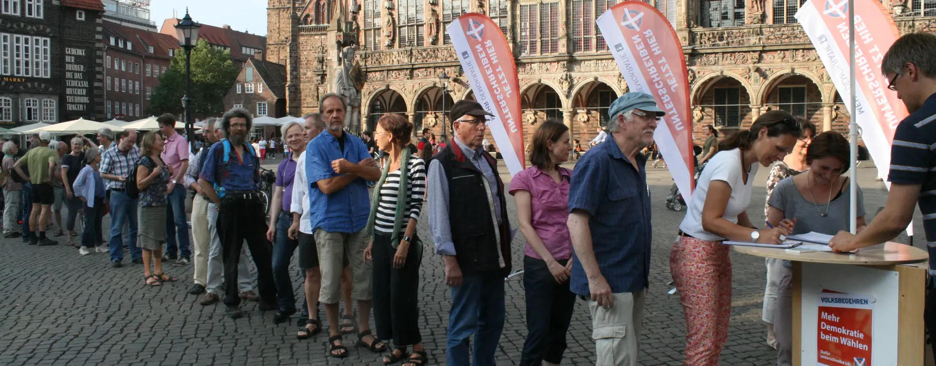 Auf einem Marktplatz sieht man eine lange Schlange, welche bei einem Volksbegehren unterschrieben will.