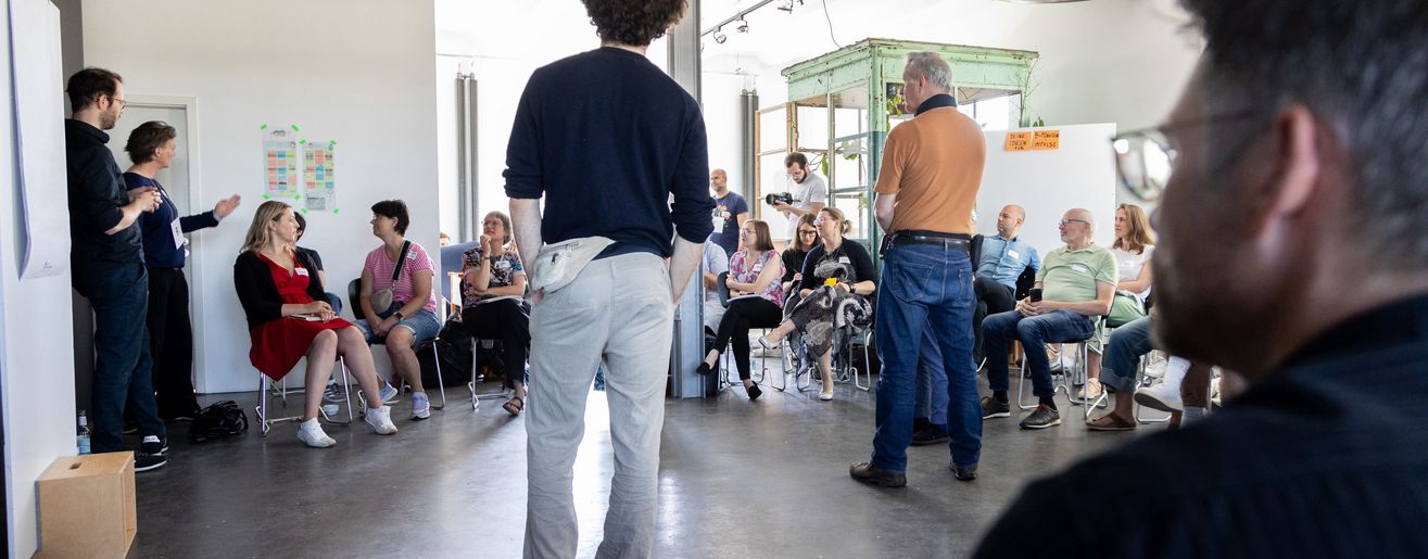 Szene einer demokratischen Systemaufstellung, mehrere Personen stehen stellvertretend für gesellschaftliche Gruppen und Institutionen in einem Raum, vor Publikum.