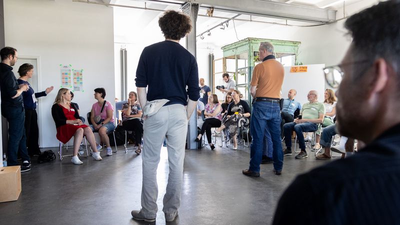 Szene einer demokratischen Systemaufstellung, mehrere Personen stehen stellvertretend für gesellschaftliche Gruppen und Institutionen in einem Raum, vor Publikum.