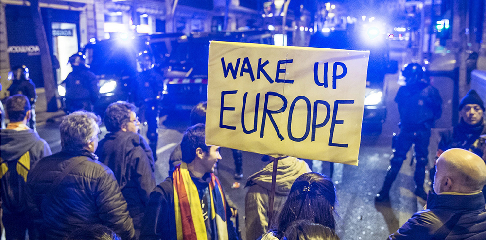 Auf einem Protest, stehen Menschen vor Polizisten mit einem Schild, wo drauf steht "Wake up Europe".