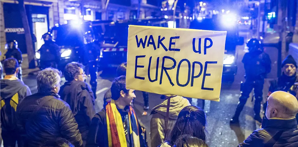 Auf einem Protest, stehen Menschen vor Polizisten mit einem Schild, wo drauf steht "Wake up Europe".