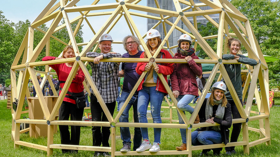 Menschen mit Bauarbeiterhelmen stehen in einer Kuppel aus Holzbalken