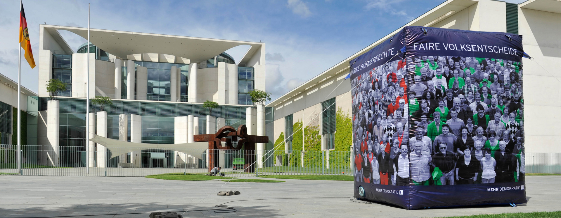 Überschrift: Direkte Demokratie/Petitionsrecht. Das Bild zeigt das Kanzleramt mit einem Würfel davor, von den ungefähren Maßen zwei mal drei Metern. Die Aufschrift lautet "Faire Volksentscheide" und auf der anderen Seite steht Starke Bürgerrechte. Dieser Würfel hat das Logo von "Mehr Demokratie"