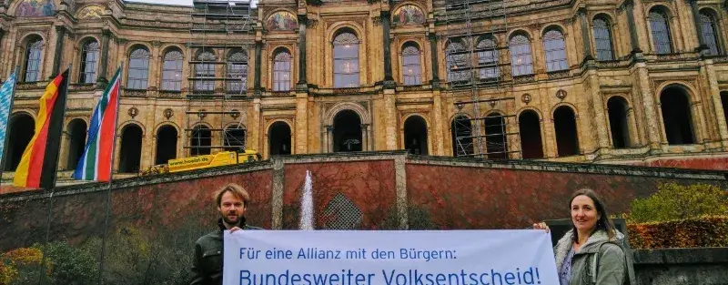 Ein Bild vor dem bayrischen Landtag, von zwei Menschen mit einem Transparent und der Aufschrift "Bundesweiter Volksentscheid".