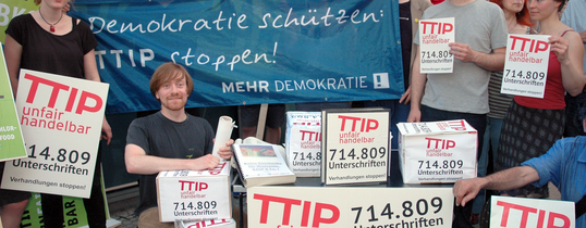 (Übergabe von 715.000 Unterschriften an die deutschen Spitzenkandidierenden für das Europaparlament | Quelle: Mehr Demokratie e.V. | Lizenz: CC BY-NC 2.0) Auf dem Bild ist eine Unterschriftensammlung gegen TTIP ist zu sehen und Menschen, die zeigen, dass schon 712.809 Unterschriften gesammelt wurden.