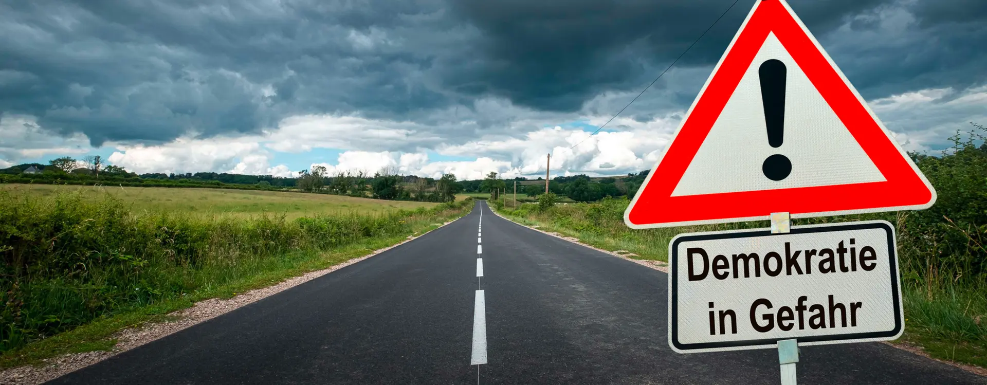 Man sieht eine Landstraße und es zeiht ein Gewitter auf, auch wenn ganz am Ende Teile eines blauen Himmels zu sehen sind. Im Fokus des Bildes ist aber ein Straßenschild mit einem Ausrufezeichen darin und darunter eine Schild Ergänzung mit den Worten "Demokratie in Gefahr".