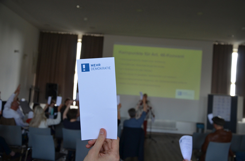 Ein länglicher Zettel wird hochgehalten mit dem Logo Mehr Demokratie. Im Hintergrund wird ein Vortrag gehalten und alle Teilnehmer:Innnen sitzen und halten ebenfalls den selben Zettel hoch.