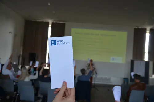 Ein länglicher Zettel wird hochgehalten mit dem Logo Mehr Demokratie. Im Hintergrund wird ein Vortrag gehalten und alle Teilnehmer:Innnen sitzen und halten ebenfalls den selben Zettel hoch.