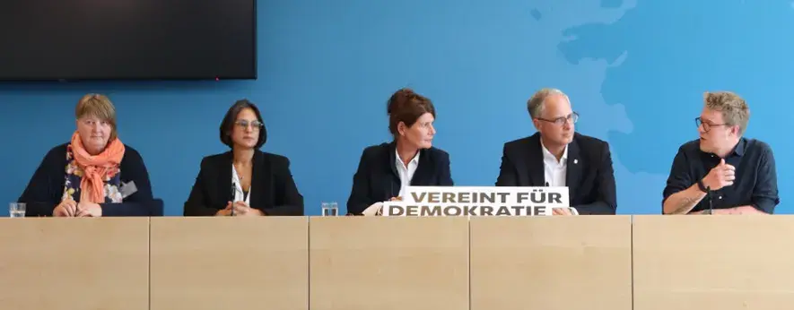 Das Bild ist von der Aufmachung an die Bundespressekonferenz angelehnt und zwei Personen halten ein Schild hoch, mit dem Text "VEREIN FÜR DEMOKRATIE".