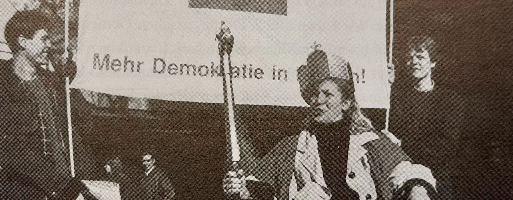 Die Bürgerin wird Königin Aktion Der Bürger wird König/Die Bürgerin wird Königin während des Volkksbegeherens Mehr Demokratie in Bayern 1995. Man sieht eine zur Königin gekrönte Bürgerin mit einem Szepter, auf einem Thron sitzend. Im Hintergrund ein Plakat mit der Aufschrift Mehr Demokratie in Bayern und dem Logo mit dem Ausrufezeichen.