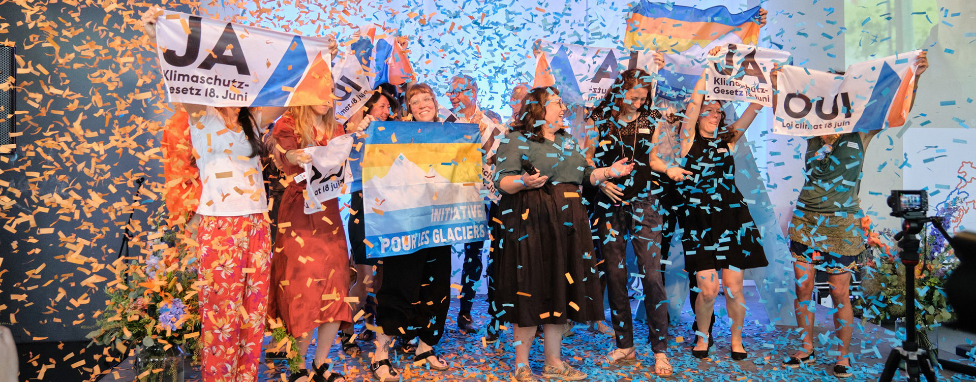 Circa 10 Menschen stehen auf einer Bühne mit Transparenten, welche die Aufschrift "JA Klimaschutzgesetz 18. Juni" haben. Von oben kommt über die Menschen ganz viel blaues und orangenes Konfetti.