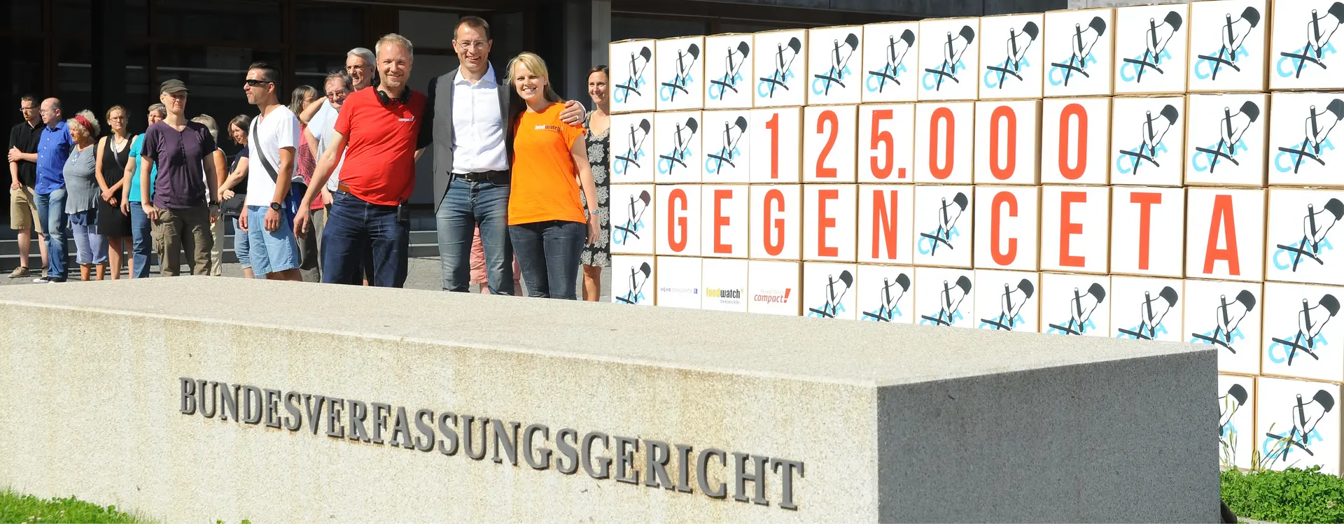 Überschrift: Handelsverträge. Es steht ein Gruppe vor dem Bundesverfassungsgericht und hält sich in den Armen. Auf einer Art Leinwand steht "125.000 GEGEN CETA". Links unten in der Ecke steht: "Mehr Demokratie" "Foodwatch" "compact!". Dass alles ist umgeben von einer Hand mit einem Stift, welche CETA durchstreicht.