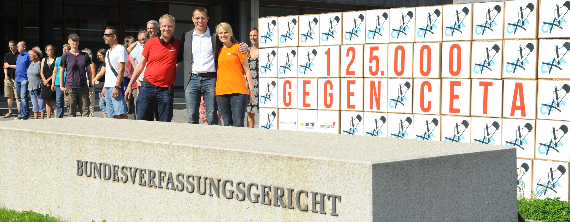 Überschrift: Handelsverträge. Es steht ein Gruppe vor dem Bundesverfassungsgericht und hält sich in den Armen. Auf einer Art Leinwand steht "125.000 GEGEN CETA". Links unten in der Ecke steht: "Mehr Demokratie" "Foodwatch" "compact!". Dass alles ist umgeben von einer Hand mit einem Stift, welche CETA durchstreicht.