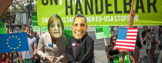 Auf dem Protestbild ist ein Transparent im Hintergrund, wo "UNFAIR HANDELBAR" steht. "FAIR" steht in weiß und der Rest in schwarz da. davor steht eine Person mit Merkel Maske und ein Mann mit Obama Maske.