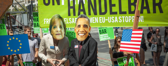 (Fotograf: Jakob Huber/Campact | Lizenz: CC BY-NC 2.0) Auf dem Protestbild ist ein Transparent im Hintergrund, wo "UNFAIR HANDELBAR" steht. "FAIR" steht in weiß und der Rest in schwarz da. davor steht eine Person mit Merkel Maske und ein Mann mit Obama Maske.