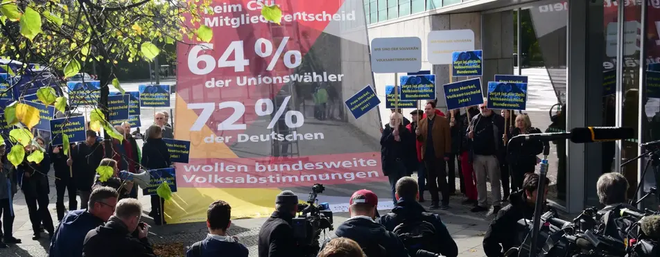 Es ist ein Protest vor einem Gebäude zu sehen mit Kameras und Transparenten.