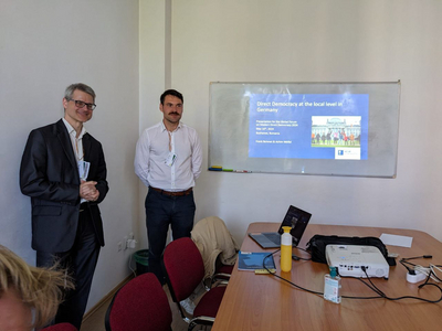 Zwei Menschen stehen vor einer Wand mit einer Präsentation an die Wand projiziert.