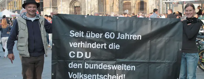 2013-08-30 Bild mit Banner Vor einer Kirche stehen zwei Menschen, welche ein schwarzes Transparent hochhalten, mit der Aufschrift "Seit über 60 Jahren verhindert die CDU den bundesweiten Volksentscheid!".