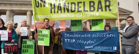 (Fotograf: Christian Mang | Lizenz: CC BY-NC-SA 2.0) Vor dem Brandenburger Tor ist eine Protestaktion gegen TTIP. Auf einem Transparent steht "UNFAIR HANDELBAR, Absatz FREIHANDELSABKOMMEN EU- USA STOPPEN!".