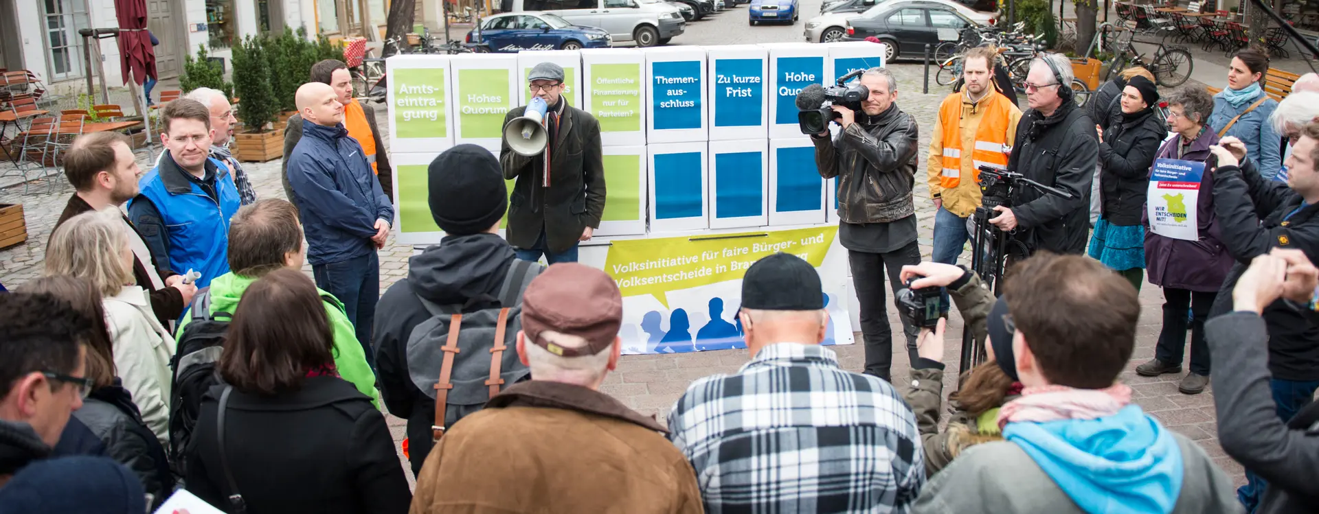 Ein Bild von einer Art Kundgebung mit Fernseh-Team und Fotografen und interessierten von Mehr Demokratie zum "Themenausschluss" und "Amtseintragung".