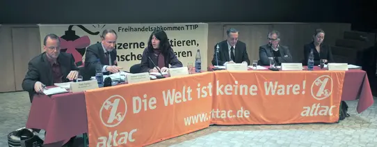 (Quelle: Mehr Demokratie e.V. | Lizenz: CC BY-NC 2.0) Auf einem Podium sitzen sechs Menschen vor einem Tisch mit dem Transparent davor gespannt, welches den Text "Die Welt ist keine Ware" trägt.