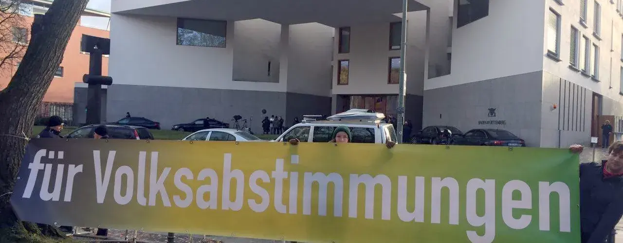 Vor einem Haus stehen drei Menschen mit einem Transparent, mit der Aufschrift "für Volksabstimmungen".