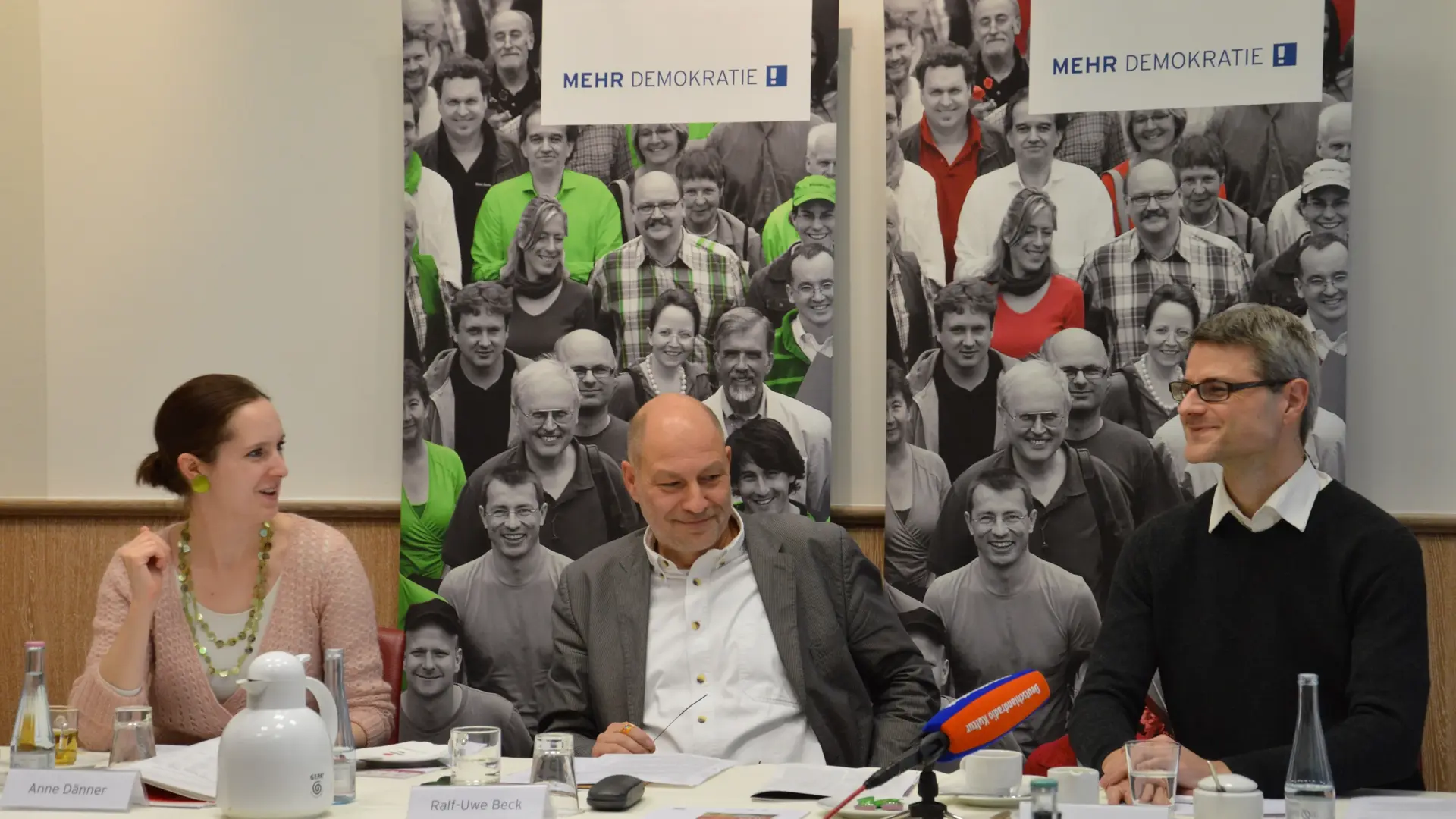 Auf einer Konferenz sieht man Ralf-Uwe Beck mit noch zwei anderen Menschen.