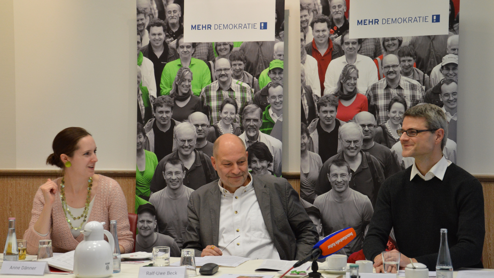 Auf einer Konferenz sieht man Ralf-Uwe Beck mit noch zwei anderen Menschen.