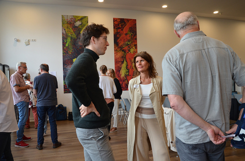 Auf dem Bild stehen drei Menschen im Kreis und diskutieren. Im Hintergrund sieht man andere Gruppen im Raum, die ebenfalls diskutieren.