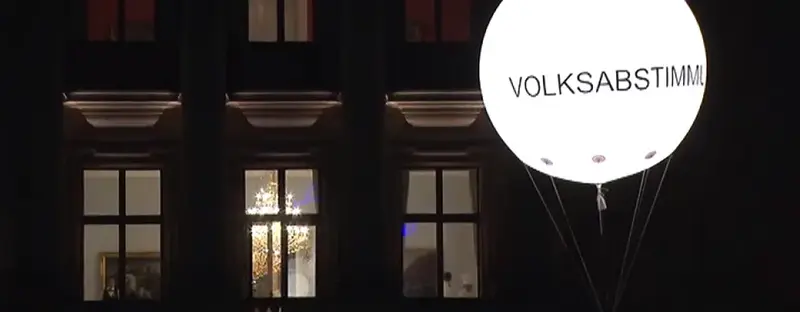 Nachts vor einem Gebäude schwebt ein leuchtender Luftballon, mit der Aufschrift "VOLKSABSTIMMUNG".
