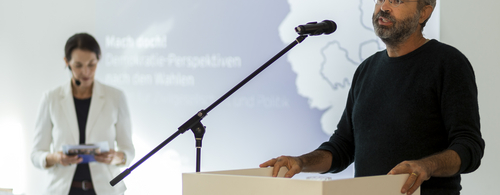 Ein Redner steht am Rednerpult, während eine Frau hinter im steht.