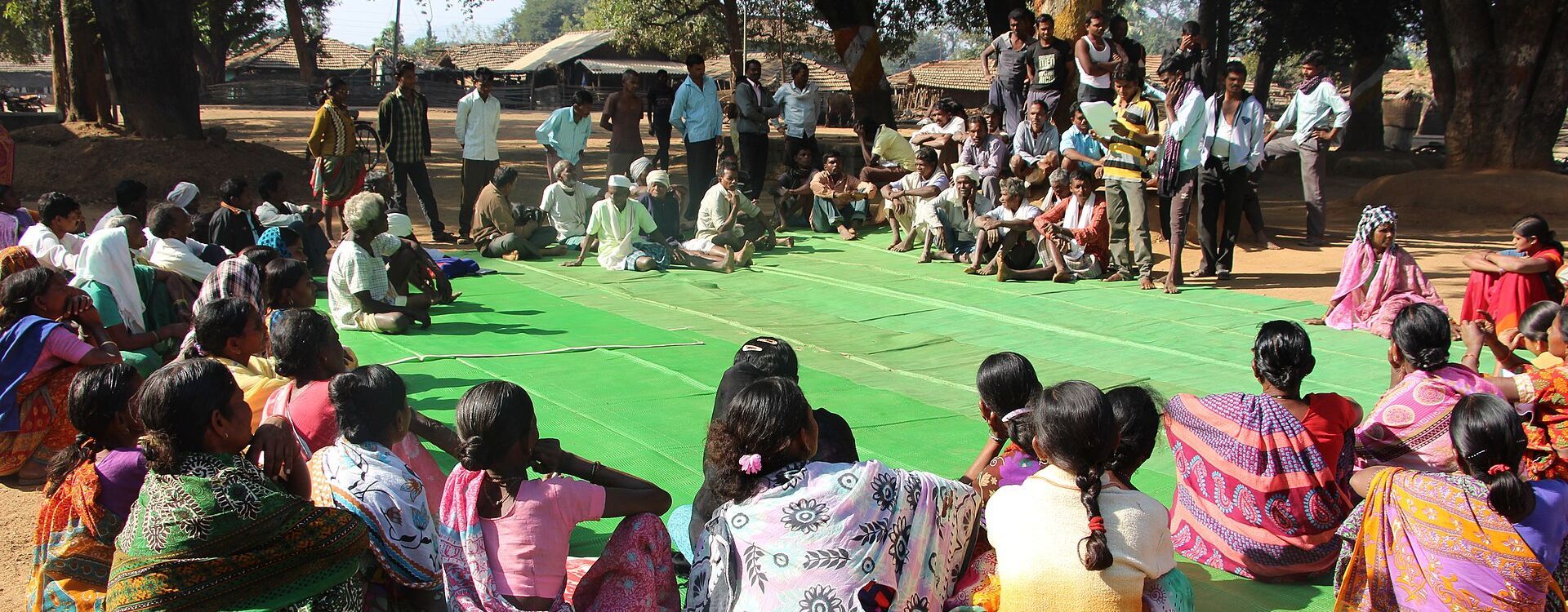 Bewohnerinnen und Bewohner des indischen Dorfes Mendha Lekha sitzen im Freien unter Bäumen zu einer Gram Sabha zusammen. In der Dorfversammlung beraten die Bürgerinnen und Bürger gemeinschaftlich über Angelegenheiten ihres Dorfes