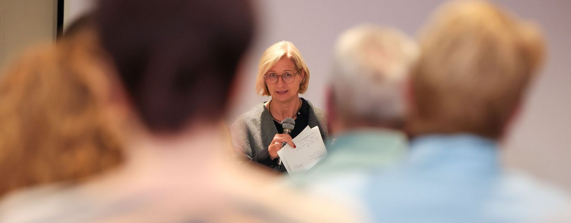 Die saarländische Ministerpräsidentin Anke Rehlinger spricht bei einem Bürgerdialog zum Strukturwandel im Landkreis Merzig-Wadern mit einem Mikrofon zu den Teilnehmerinnen und Teilnehmern. Im Vordergrund sind mehrere Zuhörerinnen und Zuhörer von hinten und unscharf zu sehen; der Blick wird dadurch auf die Rednerin in der Bildmitte gelenkt.