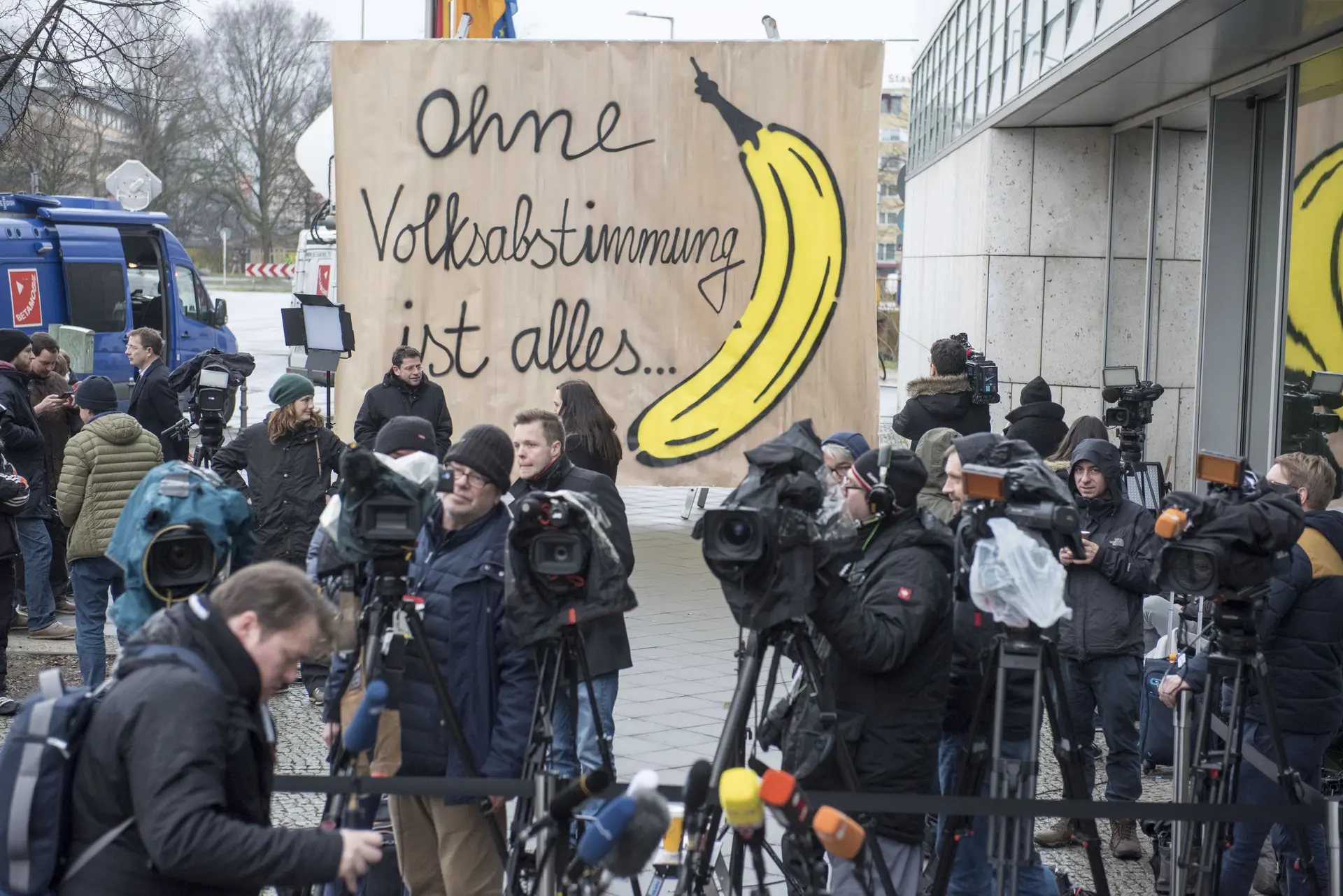 Auf dem Bild sind ganz viele Kameramänner und im Hintergrund ein Transparent mit dem Text "Ohne Volksabstimmung ist alles ..." und rechts ist eine Banane gemalt.