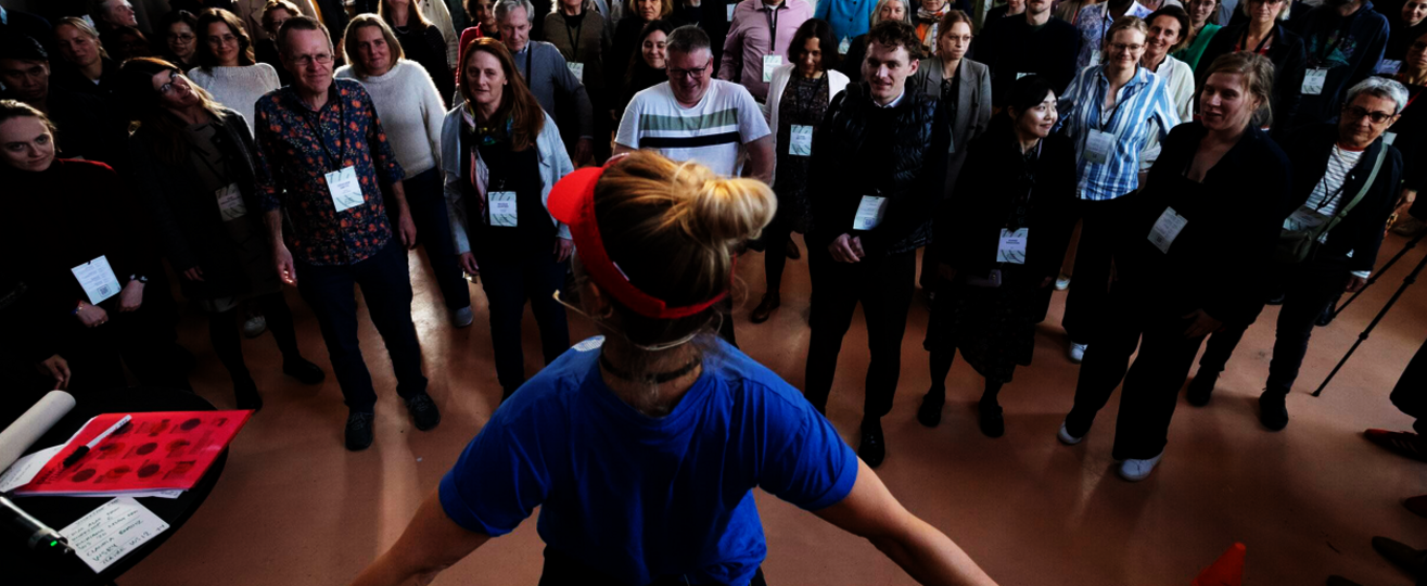 Rund 40 Menschen mit Namensschildern stehen im Kreis in einem hellen Saal; vorn leitet eine Frau mit Stirnband eine Übung zu Demokratie Fitness an.
