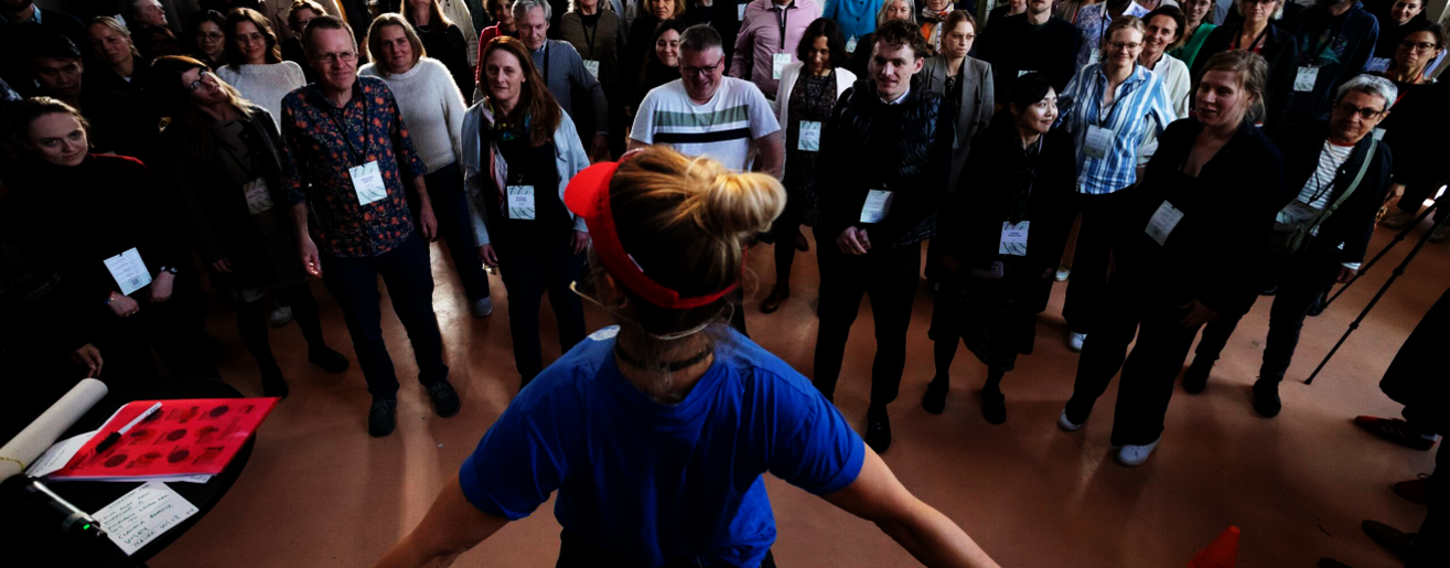Rund 40 Menschen mit Namensschildern stehen im Kreis in einem hellen Saal; vorn leitet eine Frau mit Stirnband eine Übung zu Demokratie Fitness an.