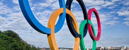 Die Olympischen Ringe stehen als Skulptur auf einer Wiese.