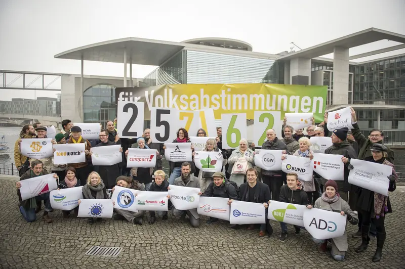 Vor einem Gebäude in Berlin stehen circa 30 Menschen, jeder hat ein Transparent in der Hand. Hinten im Bild steht "für Volksabstimmungen 257162".
