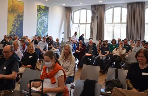 Das Bild zeigt einen fast vollen Saal mit sitzenden Menschen, die alle nach vorne gerichtet sich gerade wohl etwas anhören oder sehen, dass sieht man auf dem Bild aber nicht.