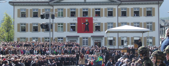 (Mit Menschen gefüllter Zaunplatz in Glarus | Foto by Cora Pfafferott | Lizenz: CC BY-NC 2.0) Auf dem Zaunplatz in Glarus stehen und sitzen circa 1.000 Menschen und drei Soldaten sind am rechten zu sehen.