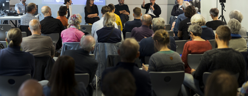 Vorne auf einer Bühne sitzen sechs Menschen und einer redet gerade zu den sitzenden Zuhörern.