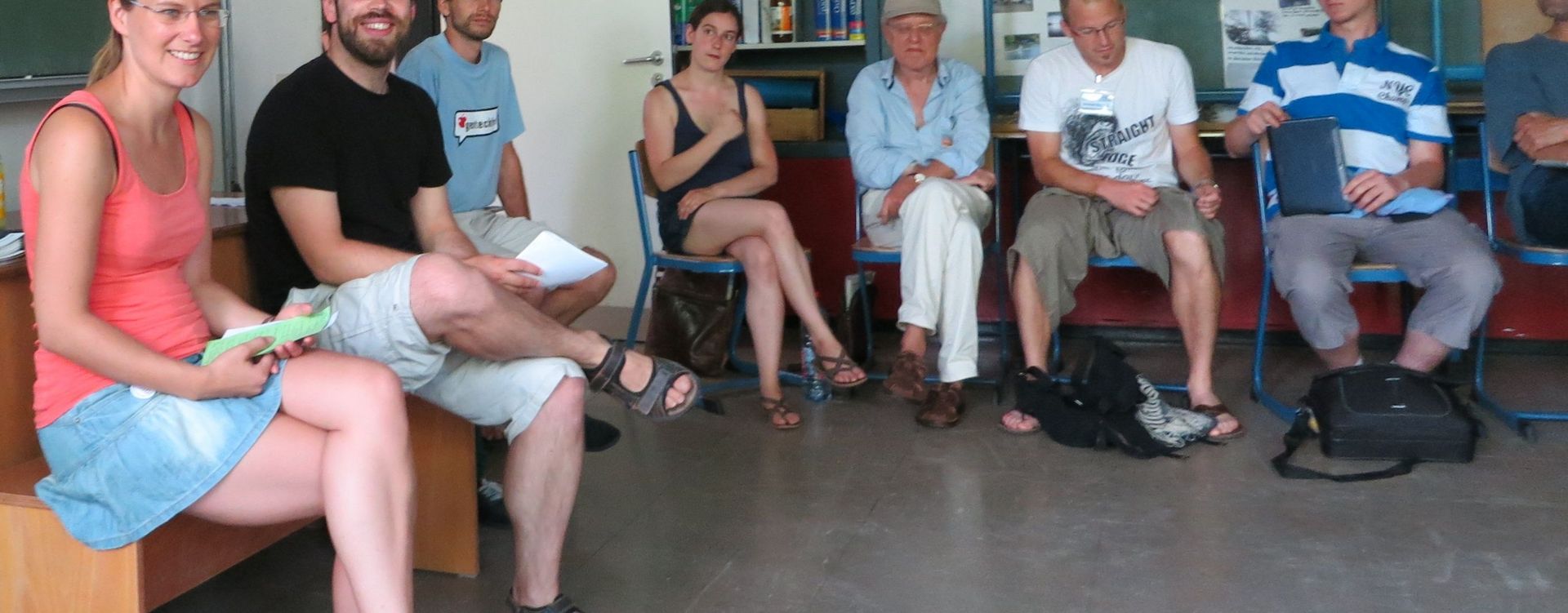 Acht erwachsene Menschen sitzen in einem Klassenzimmer und reden.