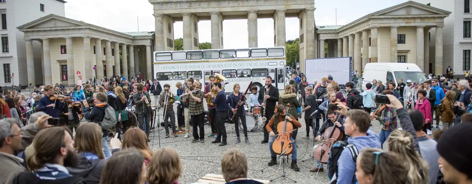 Vor dem Brandenburger Tor ist eine Menschenmenge zu sehen, ein paar der Menschen spielen Musikinstrumente. 