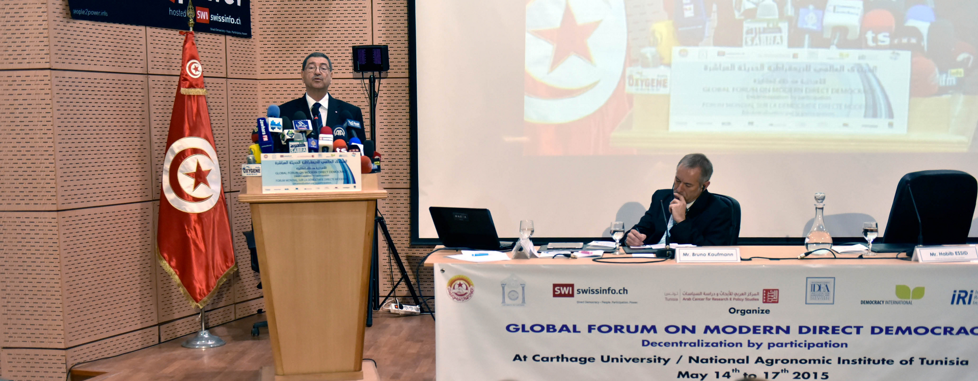 Auf dem Bild ist ein Rednerpult mit Redner und hinter ihm steht eine türkische Flagge, ein Projektor projieziert das Bild vom Redner rechts neben ihn an die Wand.