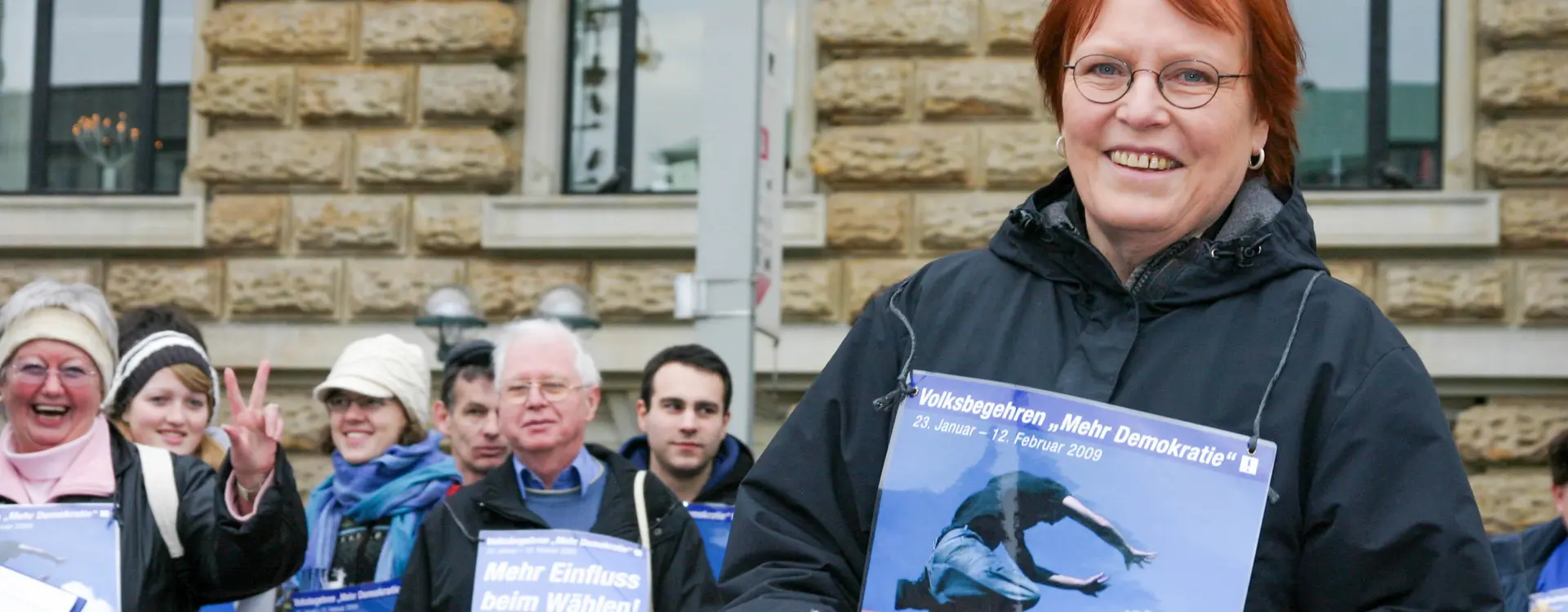 Es sind mehrere Menschen zu sehen, die alle der Kamera zugewandt sind. Die Frau im Vordergrund hat ein Plakat um der Hals, wodrauf steht: Volksbegehren "Mehr Demokratie" 23. Januar - 12. Februar 2009