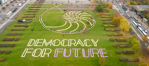 Man sieht die Wiese vor dem Bundestag mit der Aufschrift "Democracy for Future", dahinter sind Menschen auf den Umrissen eines Schneckengehäuses zu sehen.