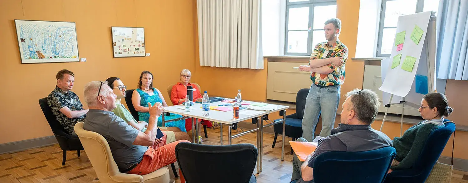 Um einen Tischen sitzen sieben Personen und eine steht vor einer Tafel und es wird augenscheinlich diskutiert.